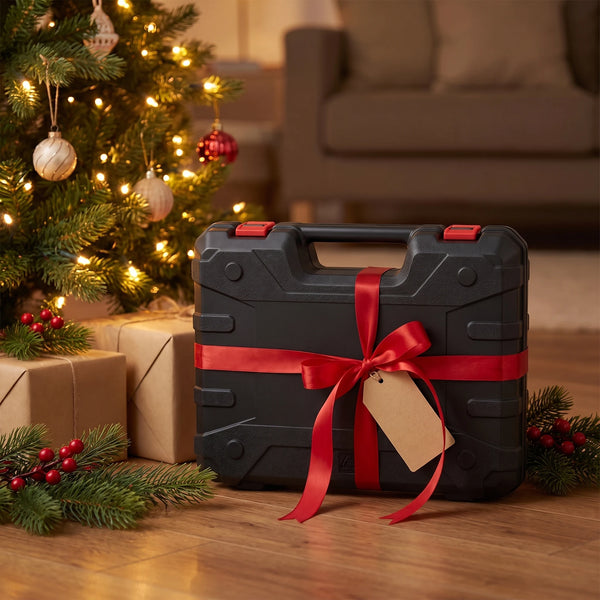 A rigid black hard case with two red latches wrapped in a loose red ribbon and a plain bow beside a lit Christmas tree, a blank kraft tag on the ribbon and wrapped boxes and pine sprigs on the floor